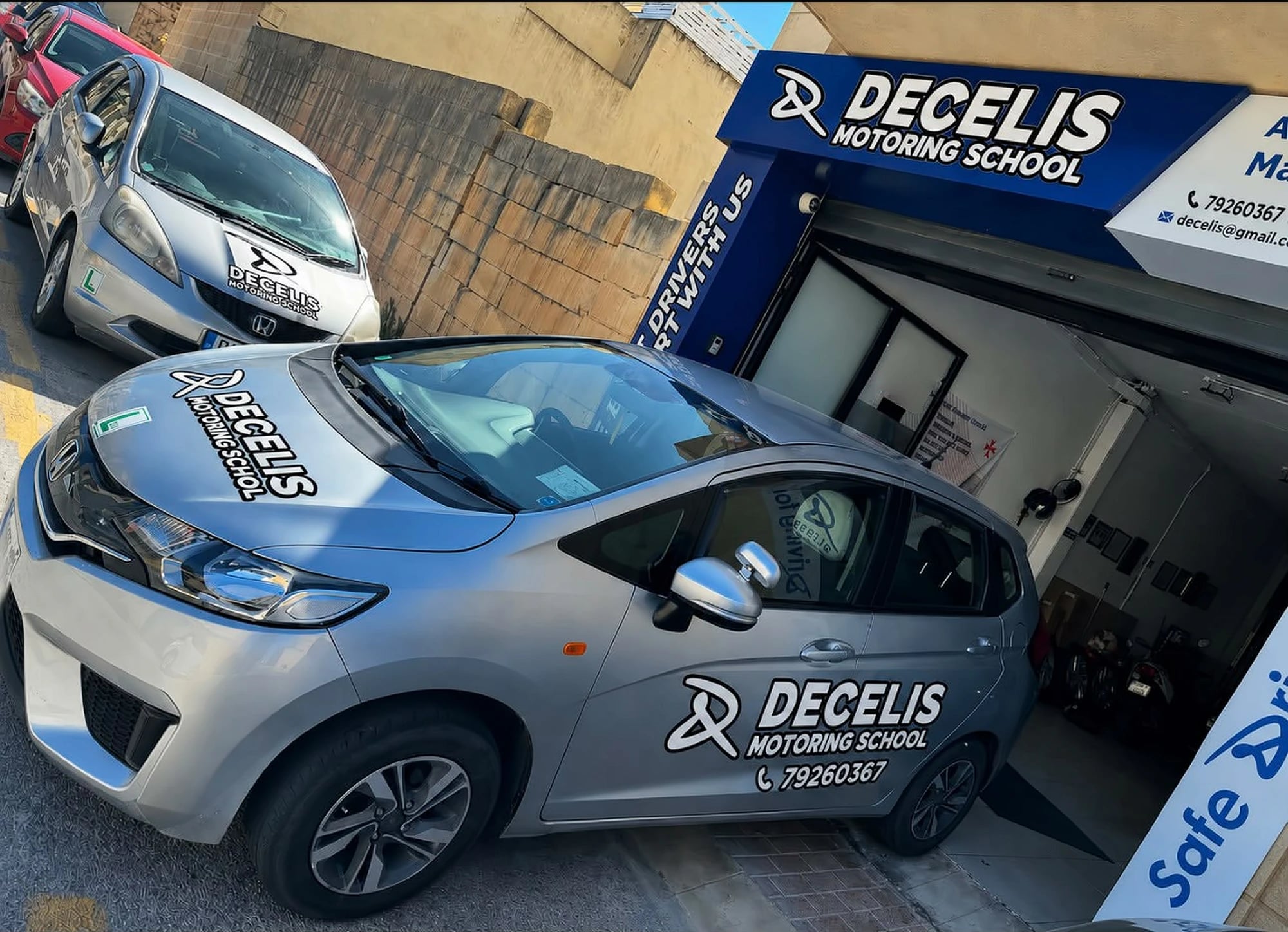 Our silver school Honda Jazz in front of the office in Żurrieq, DECELIS MOTORING SCHOOL and 79260367 on its bonnet and door, under the navy DECELIS MOTORING SCHOOL sign