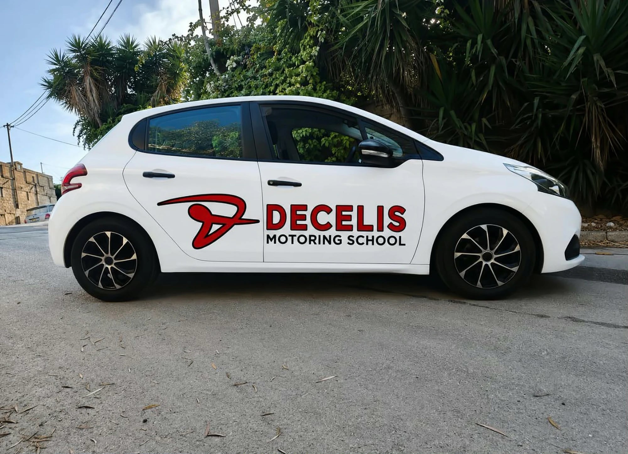 Our white Peugeot 208 with DECELIS MOTORING SCHOOL in red and black on its doors, parked by palm trees